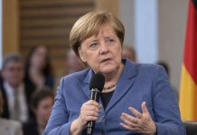 Merkel podważa wierność AfD konstytucji. Powód? Partia chce zlikwidować media publiczne! Angela Merkel z mikrofonem podczas ilustracyjnego spotkania, z flagą Niemiec w tle