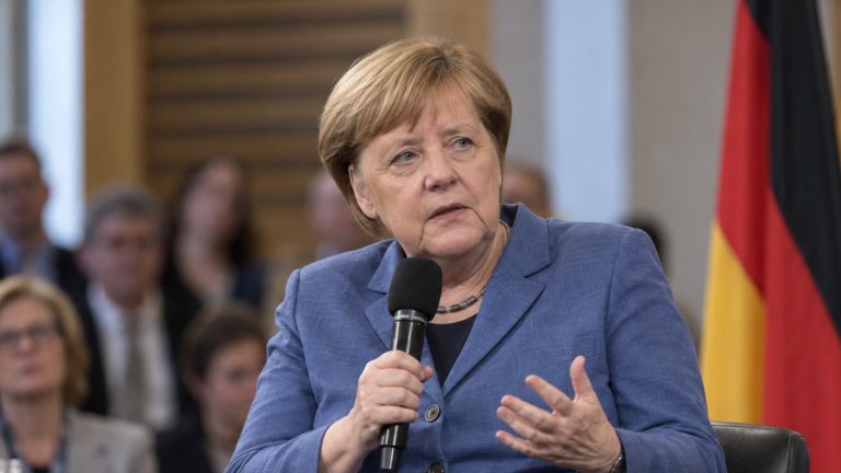Angela Merkel z mikrofonem podczas ilustracyjnego spotkania, z flagą Niemiec w tle