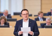 Polityk CDU odczytał w parlamencie rzekomy mail AfD z pochwałą Hitlera! Poseł zaprzecza: „To fałszerstwo” Ilustracja AI: polityk CDU Bastian Schneider odczytuje dokument przy mikrofonie w symbolicznej sali obrad.