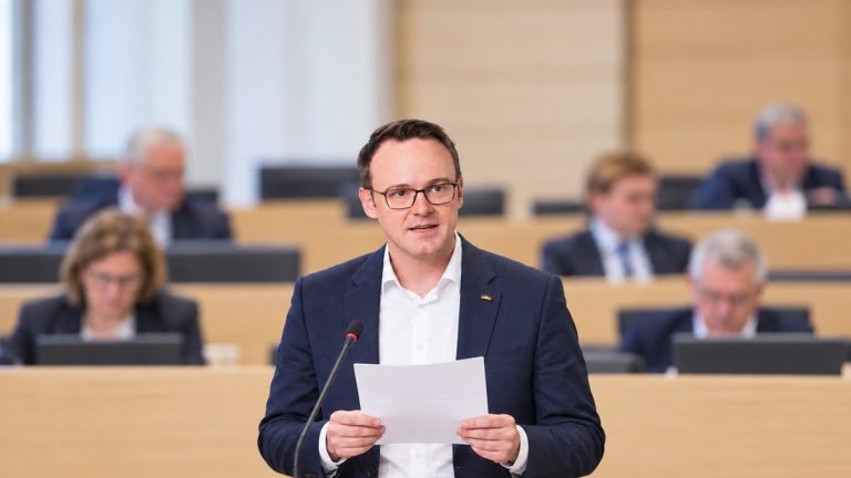 Ilustracja AI: polityk CDU Bastian Schneider odczytuje dokument przy mikrofonie w symbolicznej sali obrad.