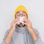 Closeup portrait of sick handsome man wearing grey sweater, yellow hat and spectacles, blowing nose into handkerchief. Male have flu, virus or allergy respiratory. Healthy, medicine and people concept