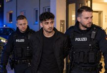 Niemcy: 17-latka zgwałcona w klatce schodowej! Policja zatrzymała 18-letniego Syryjczyka Ilustracja AI: fikcyjny dorosły mężczyzna eskortowany przez niemieckich policjantów przy wejściu do budynku mieszkalnego.
