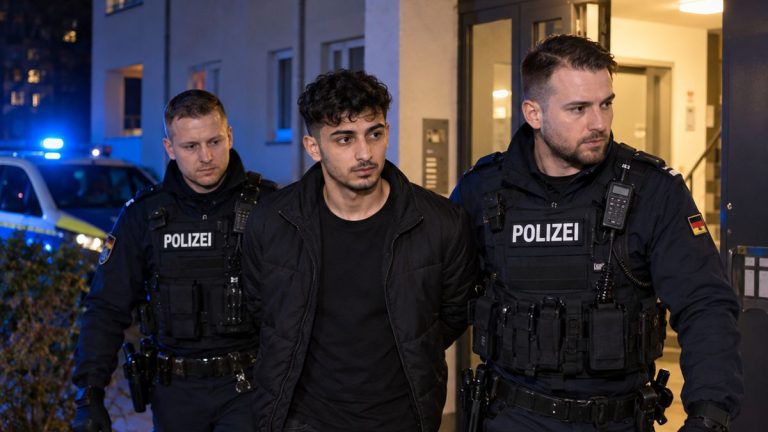 Ilustracja AI: fikcyjny dorosły mężczyzna eskortowany przez niemieckich policjantów przy wejściu do budynku mieszkalnego.