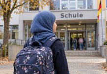 Zakaz chust dla dziewczynek w niemieckich szkołach i przedszkolach? Kobiety z CDU chcą zbadać jego wprowadzenie! Ilustracja AI: dziewczynka w chuście z plecakiem przed niemiecką szkołą.