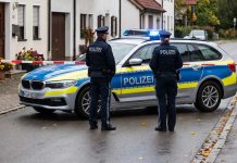 Strzały w Bawarii! Dwie osoby ranne, policja apeluje do mieszkańców: nie wychodźcie z domów! Policjanci i niemiecki radiowóz na odgrodzonej ulicy — ilustracja AI