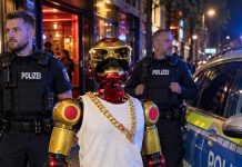 Robot molestował 16-latkę w Niemczech! Właściciel i jego kolega transmitowali zdarzenie na żywo Czerwono-złoty robot w okularach i z wąsem obok policjantów oraz radiowozu — ilustracja AI na podstawie fotografii Policji w Kolonii