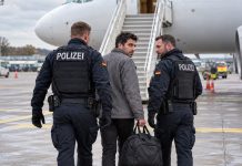 Niemcy od 2025 roku deportowały do Syrii tylko pięciu przestępców. Damaszek sprzeciwia się przymusowym powrotom! Ilustracja AI: dwaj niemieccy policjanci eskortują mężczyznę z torbą na płycie lotniska, w tle samolot i schody.